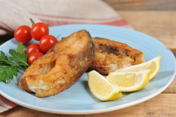 Carp steaks on a plate. Lemon slices, cherry tomatoes and parsley complete the fried fish dish. Wood background. Close-up.