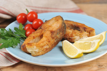 Carp steaks on a plate. Lemon slices, cherry tomatoes and parsley complete the fried fish dish. Wood background. Close-up.
