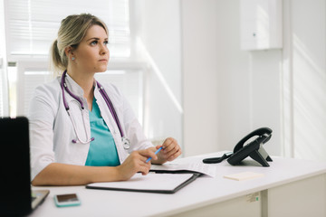 Beautiful young woman doctor sitting at the desk and writing against the background of a window. There's a laptop on the table