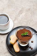 Portion of Classic tiramisu dessert in a glass cup, savoiardi cookies and espresso coffee on concrete background