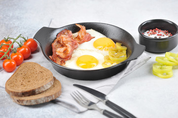 Fried eggs with bacon in a pan. Slices of dark bread, cutlery, cherry tomatoes and spices complete the composition. Close-up. Light background.