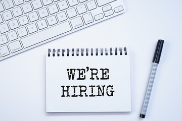 Spiral notebook with "We're hiring" text on keyboard on white background. Recruitment concept. Top view or flat lay.