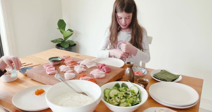 young girl puts beginner training chopsticks on before eating fresh raw sashimi while a little boy eats orange fish row - fish eggs. The raw fish is laid out on a board in the center of the table