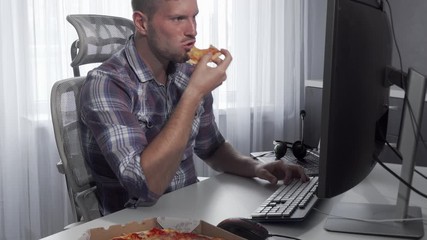 Handsome man enjoying tasty pizza while working on a computer. Young male freelancer eating pizza, sitting in front of his computer. Male student eating while studying online