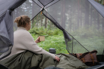 Woman covered with warm sleeping bag meets cold morning in sitting in a touristic camping tent with a cup of hot tea.