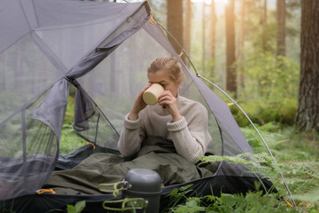 Woman covered with warm sleeping bag meets cold morning in sitting in a touristic camping tent with a cup of hot tea.