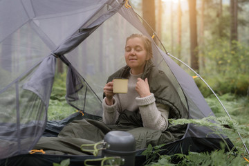 Woman covered with warm sleeping bag meets cold morning in sitting in a touristic camping tent with a cup of hot tea.