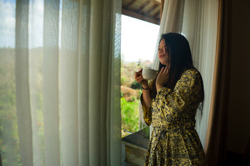 lifestyle portrait of young beautiful happy and relaxed Asian Korean woman in stylish bathrobe by luxury hotel room window having morning coffee enjoying the tropical view