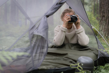 Woman covered with warm sleeping bag meets cold morning in sitting in a touristic camping tent with camera.