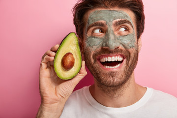 Headshot of handsome brunet man with toothy broad smile, holds slice of avocado near face, wears...