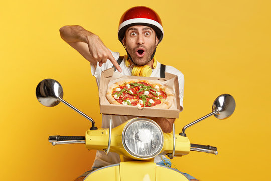 Surprised pizzaman points at delicious italian tomato pizza in cardboard box, impressed with its aromatic smell, transports tasty fast food on fast motorbike, isolated over yellow background.