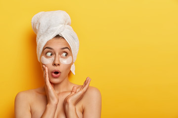 Emotional surprised Caucasian woman looks with shocked expression aside, stands bare shoulders against yellow background, has flawless soft skin, wears patches under eyes, cleans her complexion