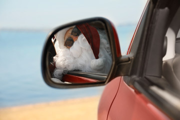 Authentic Santa Claus looking into side view mirror of car near sea