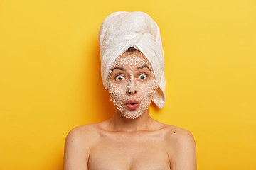 Stupefied young woman applies natural sea salt mask, reduces acnes and pimples, stares with widely opened eyes, healthy skin, cares about complexion, has spa therapy in bathroom. Hygiene and wellness