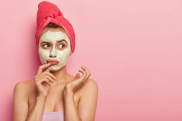 Sad upset woman wears white natural mask, has thoughtful expression, wants to have glow perfect smooth skin, applies facial clay mask, towel on head, poses over pink wall with copy space area