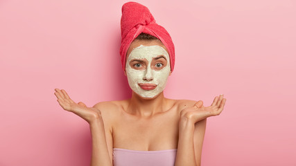 Hesitant beautiful woman with clay mask for improving skin condition, makes doubtful gesture, increases sensitivity, tries to relax after hard working week, stands against pink wall, makes decision