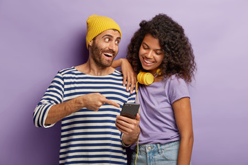 Happy guy in yellow hat and striped jumper, explains to Afro girl how to use new application at smartphone, points in display, stand closely, cannot imagine life without modern technologies.