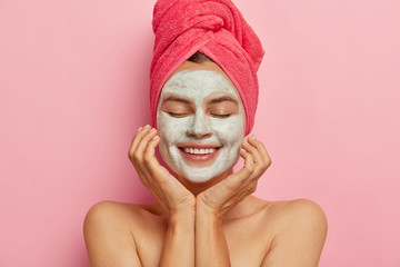 Spa girl applies clay mask on face, keeps eyes closed, touches cheeks, gets pleasure from beauty procedure, refreshes skin, smiles positively isolated on pink background. Relaxation, healthy lifestyle
