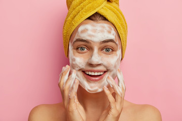 Isolated shot of pretty smiling Caucasian woman pampers face, washes skin with foaming gel, wears wrapped yellow towel on head, cares about body, isolated over pink background. Cleansing concept