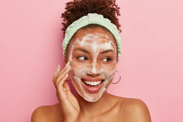 Headshot of pretty dark skinned young model touches cheek with foam, washes face with water and soap, looks gladfully aside, wears round earrings, headband, poses naked against pink background - obrazy, fototapety, plakaty