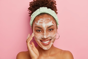 Happy dark skinned woman pampers face with bubble cleansing foam, has hygienic treatments in...