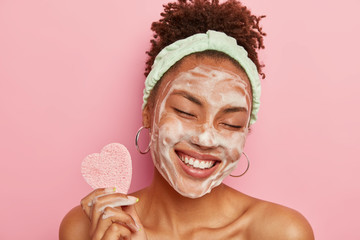 Obrazy na Plexi  Beautiful optimistic Afro American woman cleanses face with foam, refreshes skin, has well cared complexion, holds heart shaped sponge for beauty procedures, stands bare shoulders with closed eyes