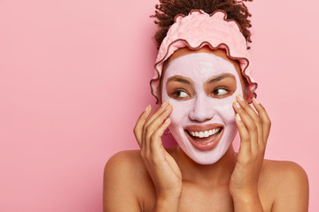 Spa and skin care concept. Pleased Afro American woman applies nourishing clay mask on face, has...