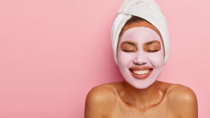 Close up portrait of young female model applies homemade facial clay mask, has white towel wrapped around head, keeps eyes shut, smiles happily, models against pink background. Beauty treatment