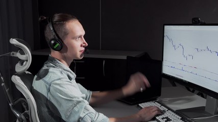 Young man using headset with microphone working online late at night. Handsome man stock trading on his computer, wearing headphones. Communication, strategy concept