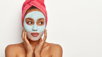 Studio shot of thoughtful dark skinned model applies facial clay mask, wrapped in towel, poses with naked shoulders, looks away against white background. People, relax and spa therapy concept