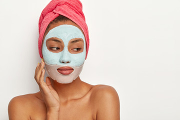 People, beauty, self care concept. Thoughtful dark skinned woman touches cheek, applies clay mask and collagen patch around lips, wears towel on head, isolated on white wall, cares about appearance