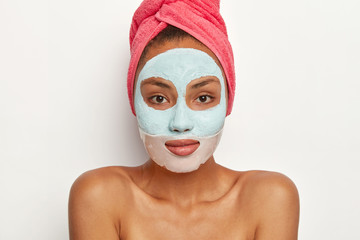 Pleasant looking female model with fresh skin, applies beauty mask, wears pink towel on head, stands with bare shoulders, looks directly at camera, poses against white wall. Skin care, spa concept