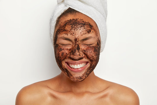 Headshot of good looking overjoyed young woman keeps eyes shut, smiles broadly, shows white perfect teeth, wears wrapped towel on head, peels face with coffee scrub, stands shirtless indoor. - Powered by Adobe