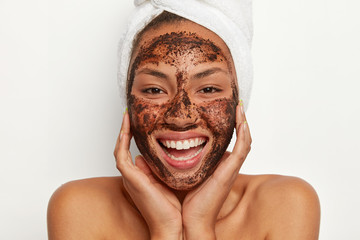 Close up portrait of happy African American woman applies coffee mask for cleansing, wants to look...