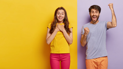 Touched pleased young European woman keeps palms on chest, gets pleasure from hearing heart warming words, overjoyed man shouts yes and clenches fists, stand together against two colored background