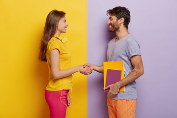 Happy groupmates meet after holidays, shake hands, agree to work together as team, stand in profile, lovely woman with headphones meets friend. Unshaven young man holds notepad talks with female