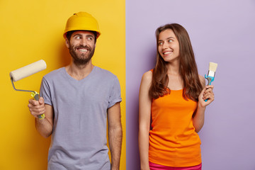Horizontal shot of happy husband and wife do repairing at home, stand with paint roller and brush, looks positively at each other, dressed casually, man wears hardhat, rejoice finally finishing work