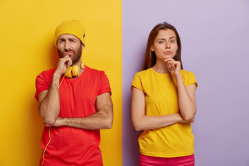 Horizontal shot of pensive millennial couple hold chins and look with displeasure at camera, wear...