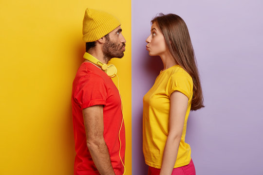 Woman and man stand in profile, keep lips folded, stare at each other, going to kiss, wear casual t shirts, headphones around neck, make grimace, pose indoor, have fun. Facial expressions concept