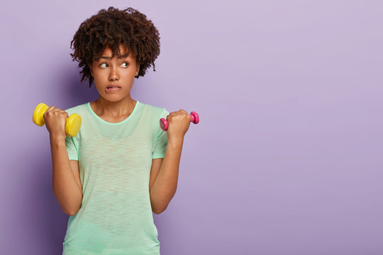 Horizontal shot of attractive curly haired woman bites lower lip, raises dumbbells, works on biceps, wears casual t shirt, focused aside, poses over purple background. Sport, healthy lifestyle concept