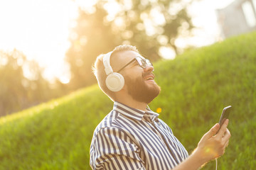 Positive young male hipster listens to your favorite online radio using smartphone headphones and high-speed wireless internet in the park on a sunny warm summer day.
