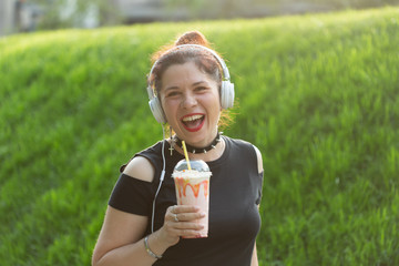 Beautiful sensual young woman listens to music with headphones and holds a milkshake in her hands while walking through the park on a sunny evening. Weekend getaway concept.