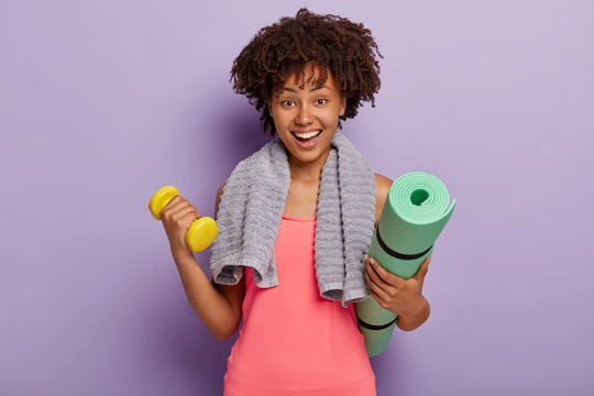 Cheerful dark skinned young Afro woman holds kareamt and dumbbell, trains muscles in gym, has happy facial expression, towel around neck, wears pink top, models indoor against purple background
