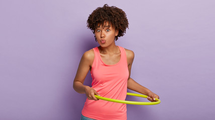 Sporty ethnic woman with curly hairstyle keeps lips rounded, rotates hula hoop, being in good shape, wears casual pink vest, has athletic body, isolated over purple background, has fitness class - obrazy, fototapety, plakaty