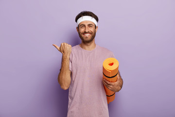 Positive unshaven guy wears white headband and purple t shirt, holds rolled up karemat, points...