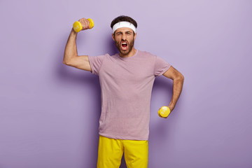 Funny sportsman raises arms with dumbbells, shouts emotionally, feels strong and sporty, dressed in purple t shirt and yellow shorts, stands indoor. Man works out in gym, does exercises. Bodybuilding