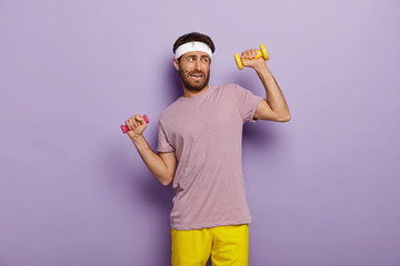 Naklejka premium Tiredness and workout concept. Displeased unshaven man raises arms with dumbbells, feels fatigue of long training, dressed in active wear, isolated over purple background, makes biceps exercises