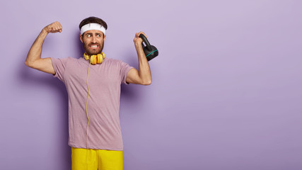 Indoor shot of handsome strong motivated man shows his biceps, raises arm, holds weight, wears white headband, casual clothes, has bodybuilding exercises, isolated on purple wall. Active lifestyle