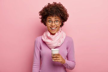 Horizontal shot of happy dark skinned woman with Afro hairdo, holds paper cup with coffee, wears purple jumper and silk scarf, poses against pink background. People, happiness, lifestyle concept