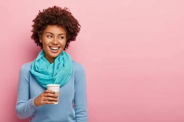 Cheerful pleasant looking woman with Afro hair drinks takeaway coffee, enjoys rest after hard working day, has pleasant talk looks aside with toothy smile dressed in blue clothes isolated on pink wall